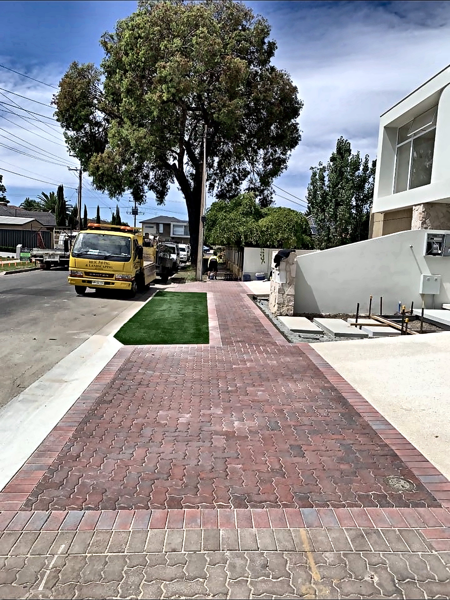Red brick paved driveway and footpath beside a new home, with a strip of lawn and a large gum tree on the street.