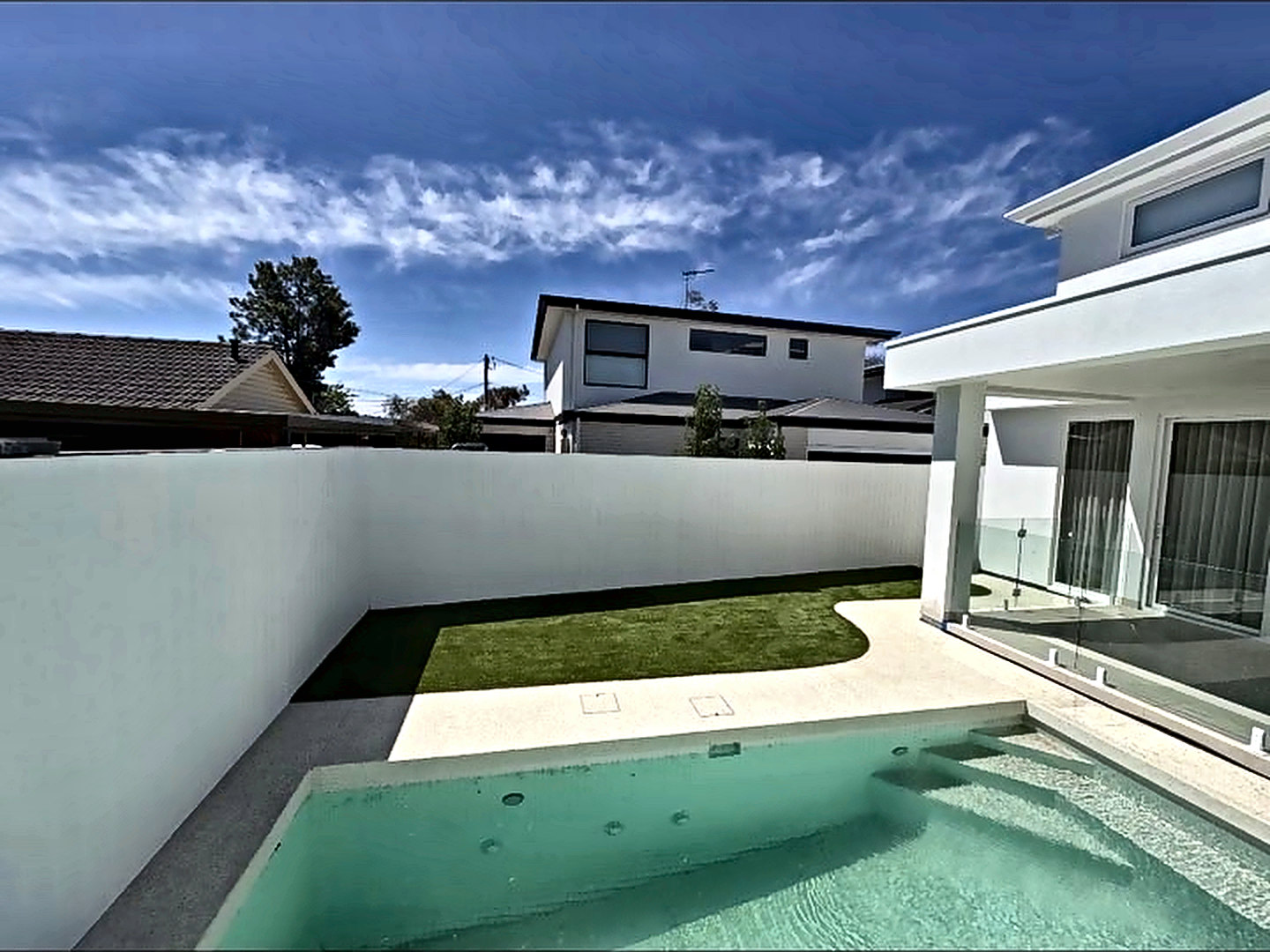 Backyard pool with pale concrete surround, synthetic lawn and rendered boundary walls.