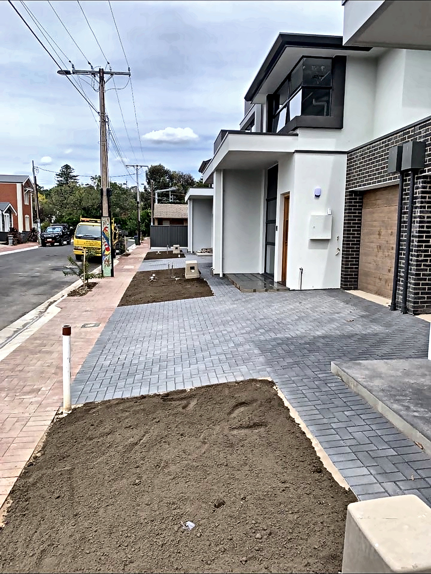 Grey paved driveway and footpath running along a row of new townhouses, with garden beds prepared for planting.